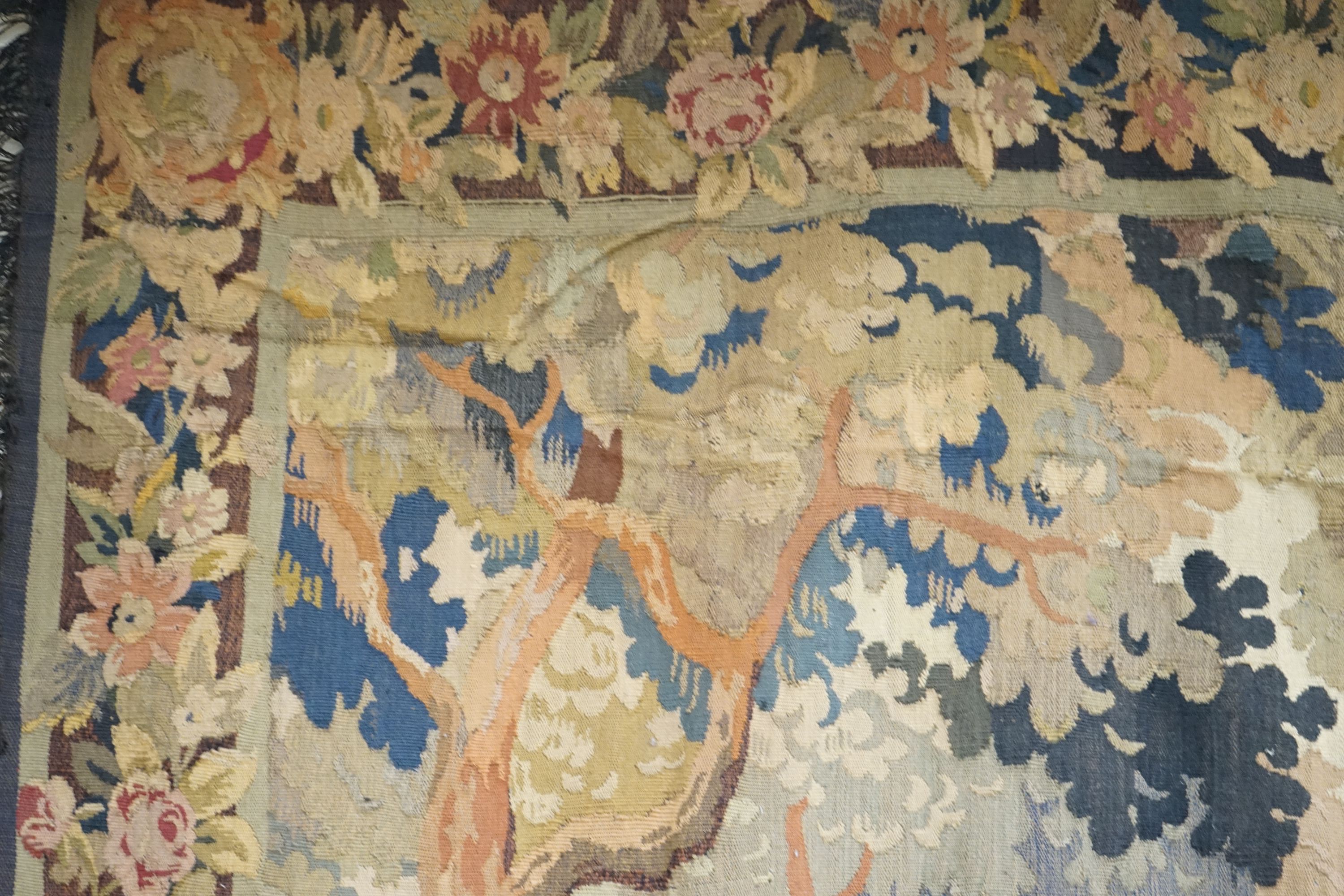 A late 19th / early 20th century French tapestry depicting a woodland scene with trees and flowers with a floral border woven in autumnal colours